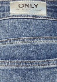 Tessuto di denim blu con una superficie liscia, caratterizzato da un'etichetta che recita "ONLY Blue Denim anno 1995." Cuciture visibili lungo le tasche.