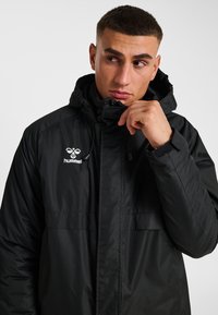 Svart huvad jacka med texturerad yta, utrustad med frontdragkedja, justerbara dragsnören och en vit logotyp på bröstet.