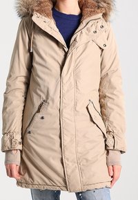 Cappotto parka beige con cappuccio foderato in pelliccia sintetica, dotato di chiusura con zip, tasche laterali e polsini a costine. Texture liscia con una vestibilità su misura.