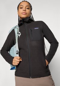 Woman with braided hair wearing a black Patagonia zip-up jacket and carrying a light blue backpack strap over her shoulder against a plain background.