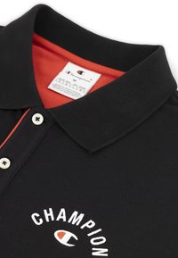 Zwarte polo met een rode binnenkraag, voorzien van een wit logo en tekst op de voorkant. Drieknoopsluiting met witte knopen.