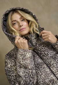 Donna con capelli biondi che aggiusta il cappuccio di un giubbotto imbottito leopardato marrone e beige su uno sfondo neutro.