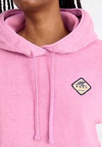 Close-up van een persoon die een roze fleece hoodie draagt met aantrekkoorden en een diamantvormige patch met een golf en de tekst "ROXY".