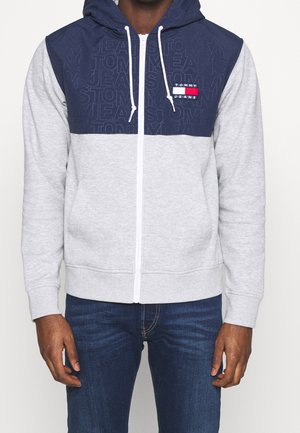 Mężczyzna w granatowej i jasno szarej bluzie z kapturem na suwak z logo Tommy Jeans na piersi, w połączeniu z ciemnoniebieskimi jeansami.