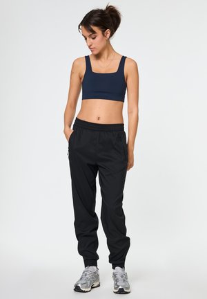 Młoda kobieta w czarnym krótkim topie, czarnych spodniach dresowych i srebrnych sportowych butach stoi z rękami w kieszeniach, patrząc w dół.