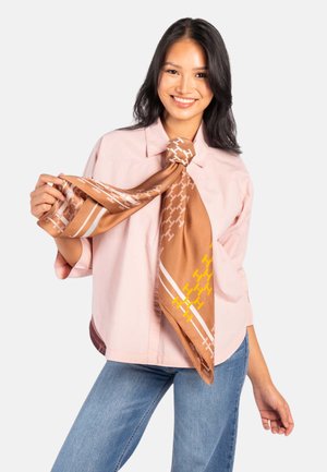 Smilende kvinne iført en lys rosa skjorte og blå jeans, som holder og viser frem et stort brunt skjerf med mønster knyttet rundt halsen.