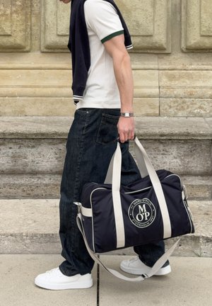 Άτομο περπατάει δίπλα σε πέτρινα σκαλοπάτια, φοράει λευκά αθλητικά παπούτσια, σκουρόχρωμα τζιν, λευκό πόλο και κουβαλάει μία ναυτική (μπλε σκούρα) τσάντα duffel με λευκούς ιμάντες και λογότυπο.