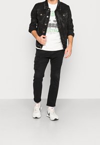 Černá denimová bunda, bílý tričko s potiskem, černé cargo kalhoty s kapsami a bílé tenisky. Klasický outfit prezentovaný na neutrálním pozadí.