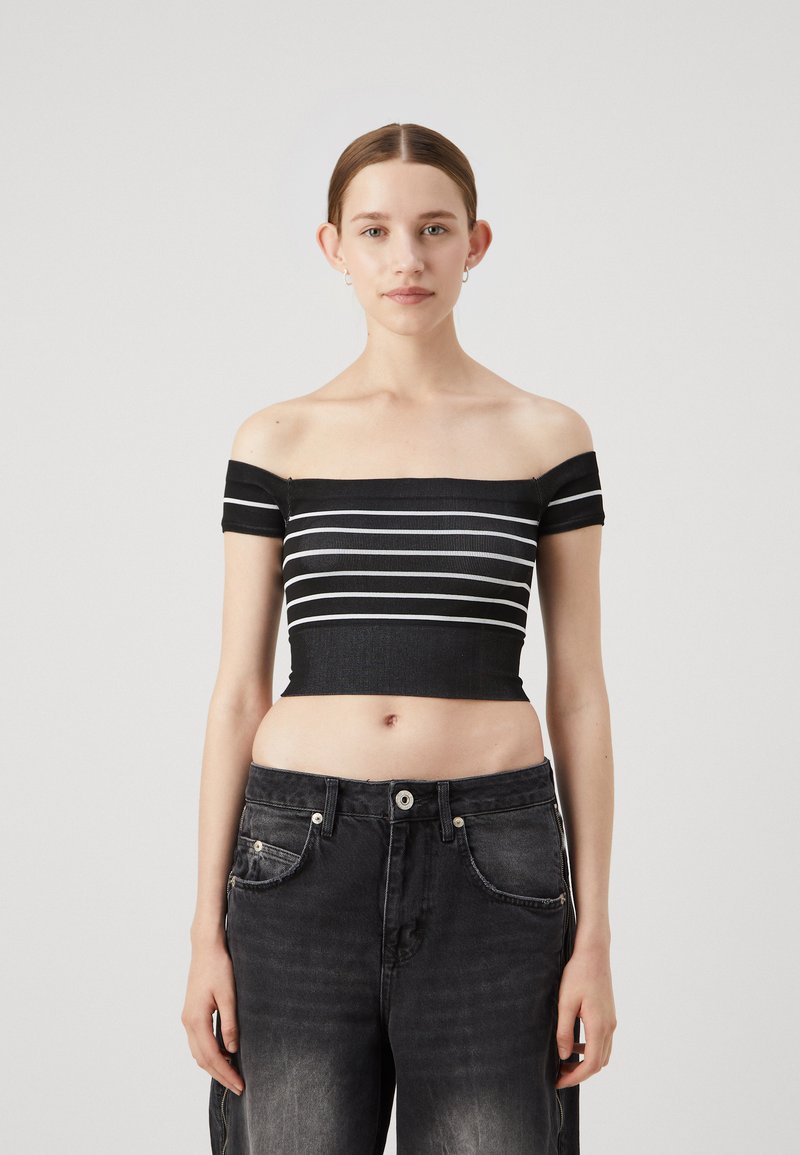 Top crop nero a spalle scoperte con strisce orizzontali bianche, texture a coste. Abbinato a jeans grigi scuri a vita alta, con cuciture visibili.