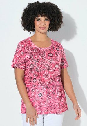 Kvinde med krøllet hår iført en kortærmet lyserød top med paisleymønster og hvide bukser, stående foran en ensfarvet lys baggrund.