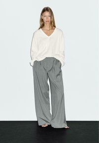 Witte lange mouwen V-hals top en grijze wijde broek, lichtgewicht stof, casual pasvorm, met plooien en zijzakken. Effen achtergrond.