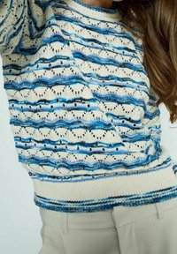 Sweter dzianinowy w kremowym kolorze z niebieskimi poziomymi paskami i wzorem ażurowym. Posiada ściągacze przy mankietach i dolnym brzegu oraz luźny krój.