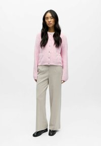 Rozā pūkaina kardigāns ar garām piedurknēm un sudraba pogām, saskaņots ar augsto vidukli gaiši pelēkiem biksēm un melniem mokasīniem. Neitrāls fons.