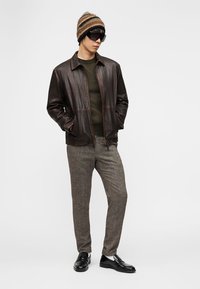 Ανδρας με καφέ δερμάτινο μπουφάν, ελιά πουλόβερ, γκρι παντελόνι, ριγέ πλεκτό σκουφί, μαύρα γυαλιά ηλίου και μαύρα μοκασίνια, στέκεται με τα χέρια στις τσέπες.