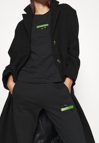 Abrigo negro de lana con botones dorados superpuesto a una camiseta negra con un gráfico verde. Pantalones negros con un parche verde en el bolsillo.