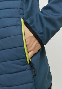 Giacca blu con texture trapuntata su un lato, dettagli neri sui polsini e una zip verde brillante a contrasto.