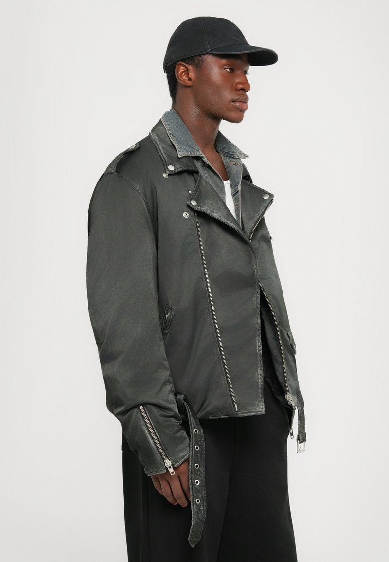 Dark gray biker jacket with zippered front and side pockets. Features a denim collar and adjustable straps at the waist. Solid black cap worn.