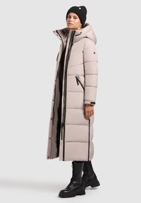 Dlhý, svetlo béžový pufkový kabát s kapucňou, s horizontálnym prešívaním, čiernymi zipsami a bočnými vreckami. Nosený na tmavé oblečenie a topánky.