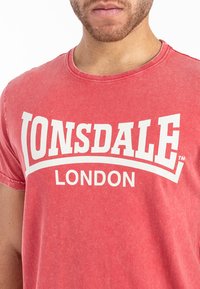 Raudona medvilninė marškinė su apvaliu kaklu, priekyje esantis bold baltas tekstas "LONSDALE LONDON" išlenktoje formoje.