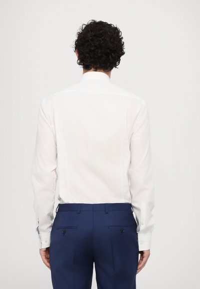 Chemise blanche à manches longues avec poignets à boutons, associée à un pantalon bleu marine ajusté. Tissu lisse avec un col classique et un détail de couture discrète au dos.