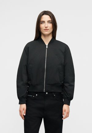 Egyenes barna hajú nő fekete zipzáras bomberdzsekit és fekete nadrágot viselve, egy egyszerű világos háttér előtt áll.