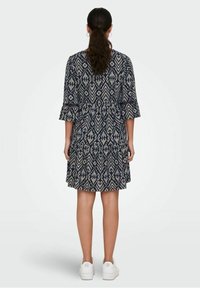 Žena s tmavými vlasy v culíku má na sobě šaty po kolena v námořnické modré barvě s vzorem a rozšířenými rukávy, bílé tenisky a stojí zády.