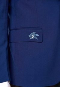 Zils žakets ar mazu izšūtu Sonic the Hedgehog logotipu uz kabatas vāka, kas parāda daļu no balto krekla piedurknes.