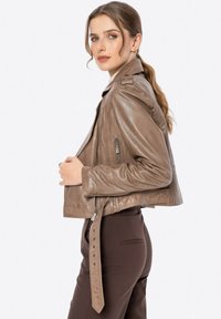 Gekürzte braune Lederjacke mit Reißverschluss-Taschen, seitlichem Gürteldetail und glatter Textur, getragen über dunkelbraunen Hosen.