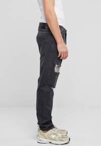 Vaqueros de mezclilla gris con un corte relajado, que presentan un desgarro desgastado en la rodilla izquierda. Combinados con zapatillas beige y una camiseta sin mangas blanca.