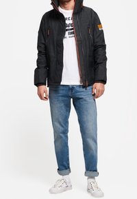 Chaqueta negra con cremallera y acentos naranjas, combinada con una camiseta gráfica blanca, jeans claros arremangados y zapatillas blancas con detalles coloridos.