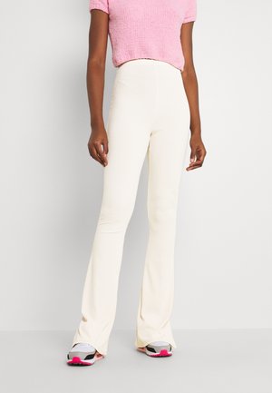 Žena v bílé žebrované kalhotách s vysokým pasem, růžovém krátkém texturovaném topu a bílých teniskách s růžovými detaily, stojící na neutrálním pozadí.