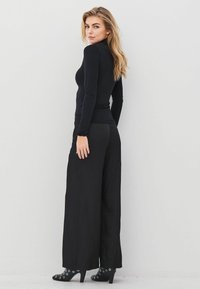 Ženska z dolgimi blond lasmi, oblečena v fitted črn top z dolgimi rokavi, široke črne hlače in črne čevlje s peto s srebrnimi luknjami.