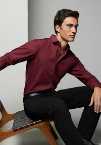 Cămașă butonată burgundy cu o textură netedă, purtată cu pantaloni negri și o curea neagră. Modelul este așezat pe un scaun din lemn cu slats.