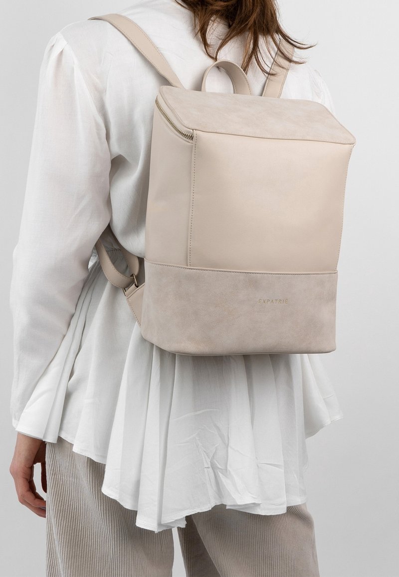 Béžový kožený batoh s hladkými a semišovými texturami, ve tvaru čtverce, s horním zipem a zlatým nápisem u spodní části.