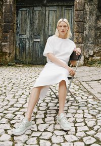 T-shirt bianca oversize e gonna lunga, abbinati a sneakers verde chiaro. Lo sfondo presenta ciottoli e una porta in legno invecchiata.