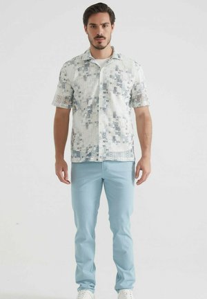 Chemise à manches courtes à boutons de couleur crème avec un motif géométrique bleu ; associée à un pantalon bleu clair et des baskets blanches.