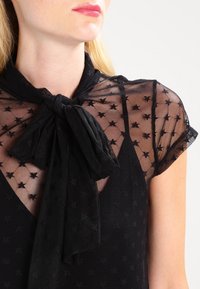 Blusa negra transparente con mangas cortas, que presenta un patrón de estrellas, un cuello con lazo y un acabado texturizado.