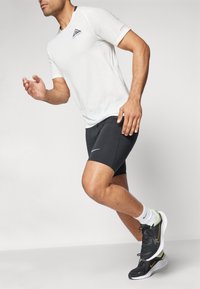 Lättviktig vit texturerad sporttröja med en svart triangel-logotyp, tillsammans med svarta åtsittande shorts och svarta löparskor med gröna detaljer.