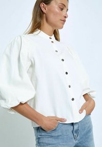 White button-up shirt with puff sleeves, made of lightweight fabric. Features silver buttons and a structured collar. Worn with blue jeans.