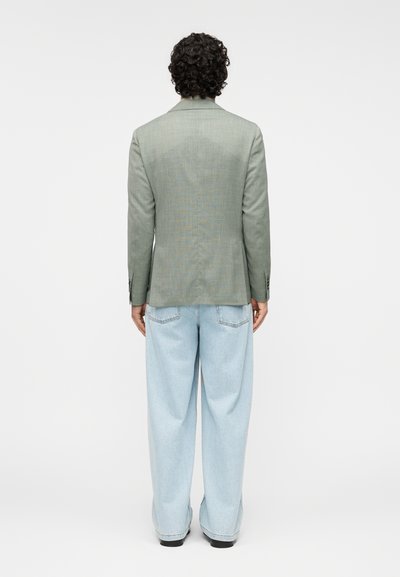 Ανοικτό πράσινο σακάκι με υφή, μονή σχισμή και σκούρα κουμπιά, σε συνδυασμό με χαλαρά ανοιχτά μπλε τζιν και μαύρα παπούτσια, από πίσω όψη.