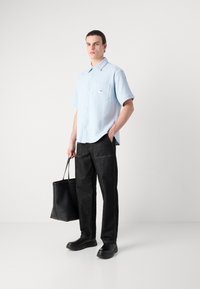 Camicia azzurro chiaro a maniche corte con finitura testurizzata, pantaloni di denim neri e una borsa tote nera. L'outfit è completato con un paio di scarpe slip-on nere.