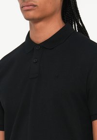 Sort polo shirt med struktureret stof, der har en tre-knaps pløs, en krave og et diskret logo broderet nær brystet.