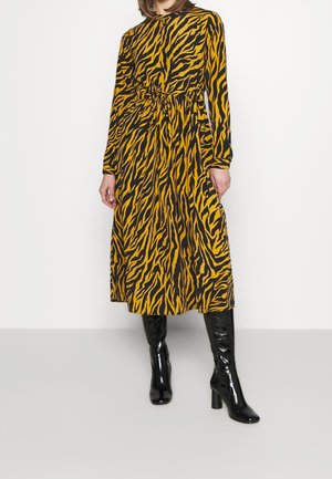 Kvinde iført en langærmet midi-kjole med gul og sort dyreprint og bundet talje, kombineret med sorte knæhøje støvler med hæl.