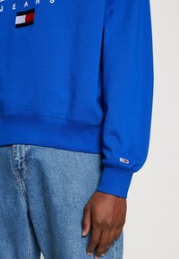 Felpa blu in cotone, con polsini a costine, un logo applicato e collo rotondo. Abbinata a jeans in denim azzurri.