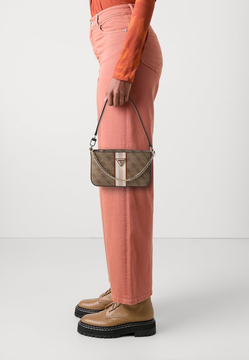 Personne portant un pantalon corail, des chaussures montantes couleur cuir clair, tenant un sac à main marron à motif avec une chaîne, sur un fond uni.