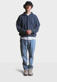 Námořnická fleecová mikina s čtvrtinovým zipem přes bílý tričko, doplněná světle modrými džíny a béžovými teniskami. Pohled zepředu, ruce sepnuté.