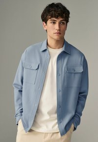 Camisa de botones azul claro con dos bolsillos en el pecho y un acabado texturizado, superpuesta sobre una camiseta blanca de cuello redondo. Fondo neutro.