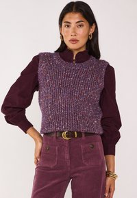 Smanicato viola lavorato a maglia con motivo strutturato, indossato sopra una camicetta viola scuro con colletto arricciato, abbinato a pantaloni di velluto a coste coordinati.