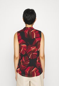 Person med kort svart hår som bär en ärmlös svart topp med stora röda och bruna lövmönster, stående mot en enfärgad vit bakgrund.