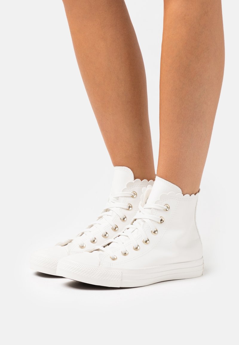 Jambes portant des baskets montantes blanches avec des bords festonnés et des lacets blancs sur un fond clair uni.